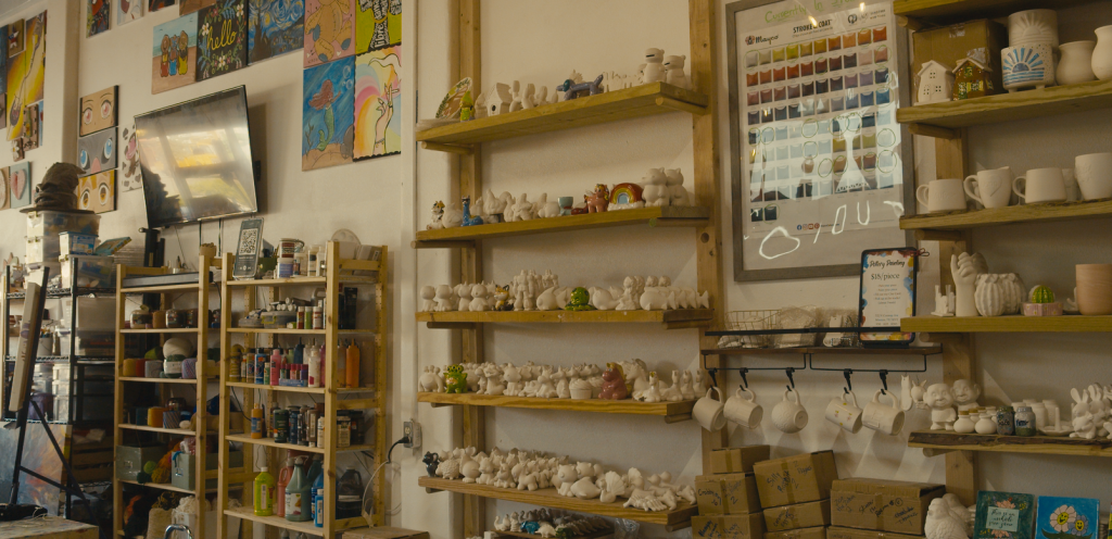 Shelves along the wall hold various ceramic pieces, yarn, and paintings.