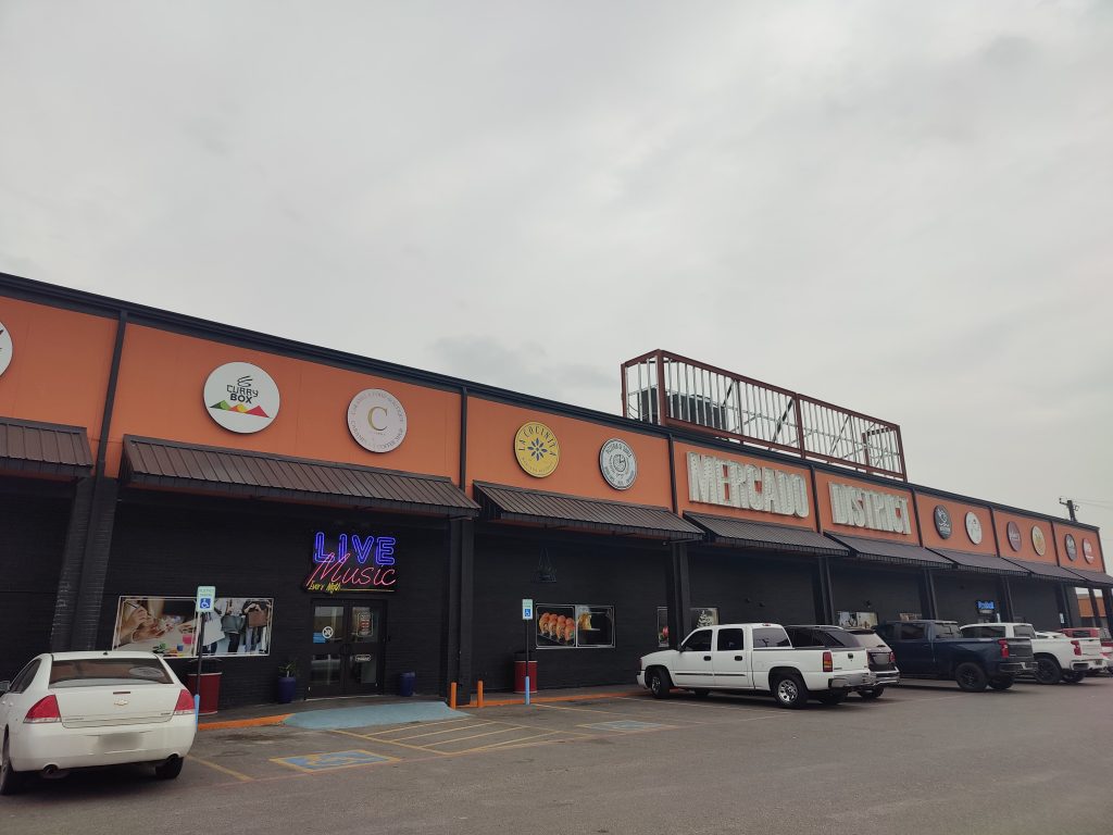 The current storefront of a warehouse has a sign that reads “Mercado District” and other store signs.