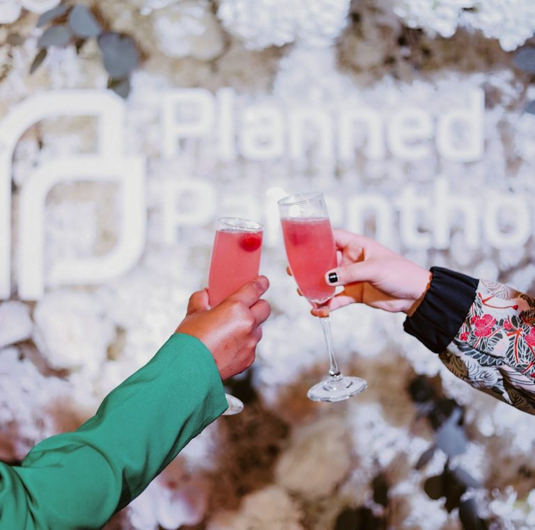 Two people cheering with champagne glasses in front of a wall decorated with flowers that read “Planned Parenthood.”