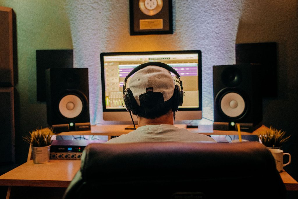 The back of a man’s head wearing a white cap and headphones. He is looking forward at a computer screen, speakers on each side of the screen.