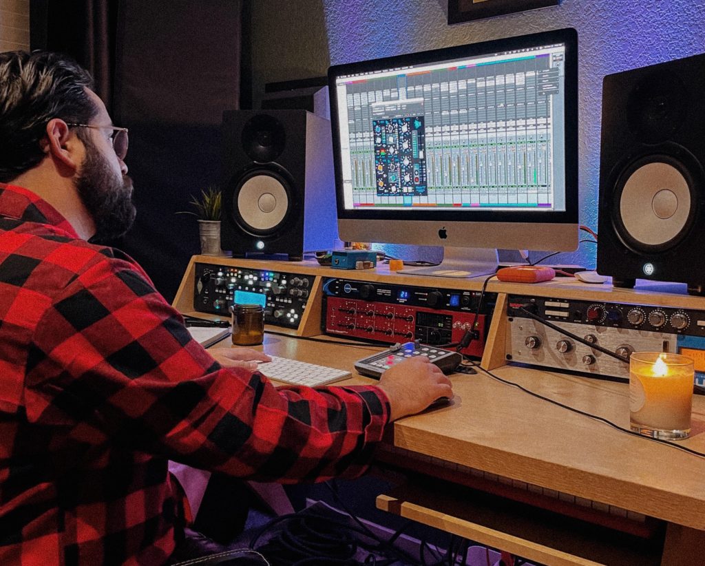 A man in a red checkered plaid shirt working on a computer, candles scattered on the desk.