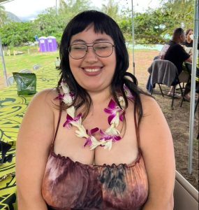 Person in a purple tye dye dress with dark hair and blunt bangs smiling, wearing glasses with her eyes closed.