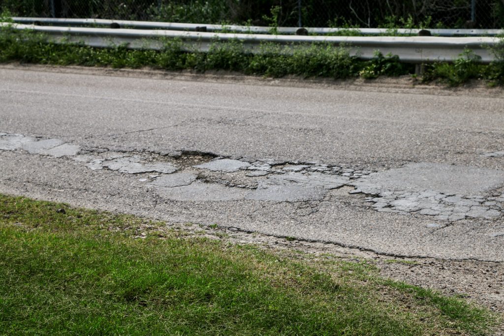 A close look at a beat-up road with a patch on the right of poorly filled and growing potholes.