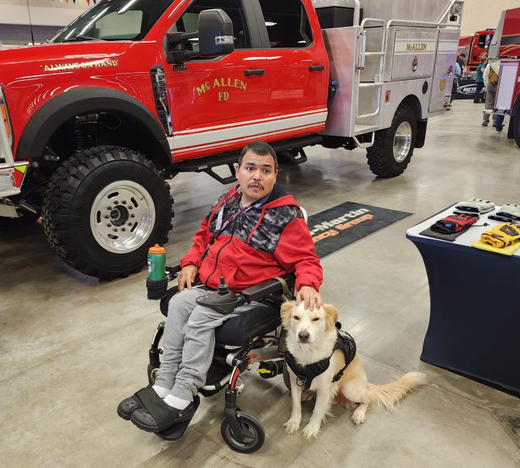 A man in a wheelchair, wearing a red jacket posing in front of a large red McAllen fire truck with his blonde Labrador mix service dog sitting next to him.