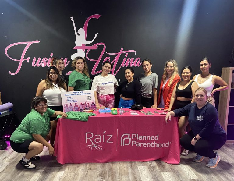 11 Creativa Resistencia workshop participants pose in front of a Pink Planned Parenthood table behind a black wall that says Fusion Latina in pink script font.