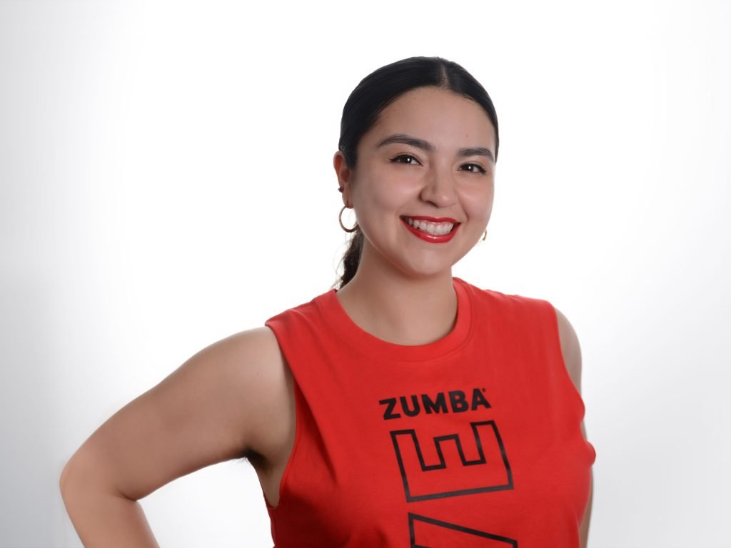 Professional headshot of Twerk instructor Alondra Vasquez, wearing a red shirt with the words Zumba. She is smiling with her hand on her waist.
