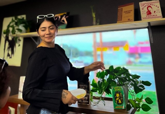 Selina Herrera, a woman in a long black shirt, with white sunglasses on her head, is touching a plant. Her gaze and smile are directed at someone speaking with her.