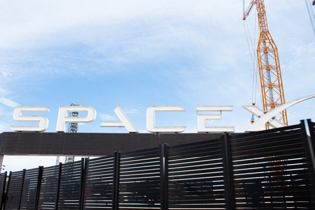 A white sign on Boca Chica Beach with the words SpaceX, bright blue clouds and metal infrastructure.