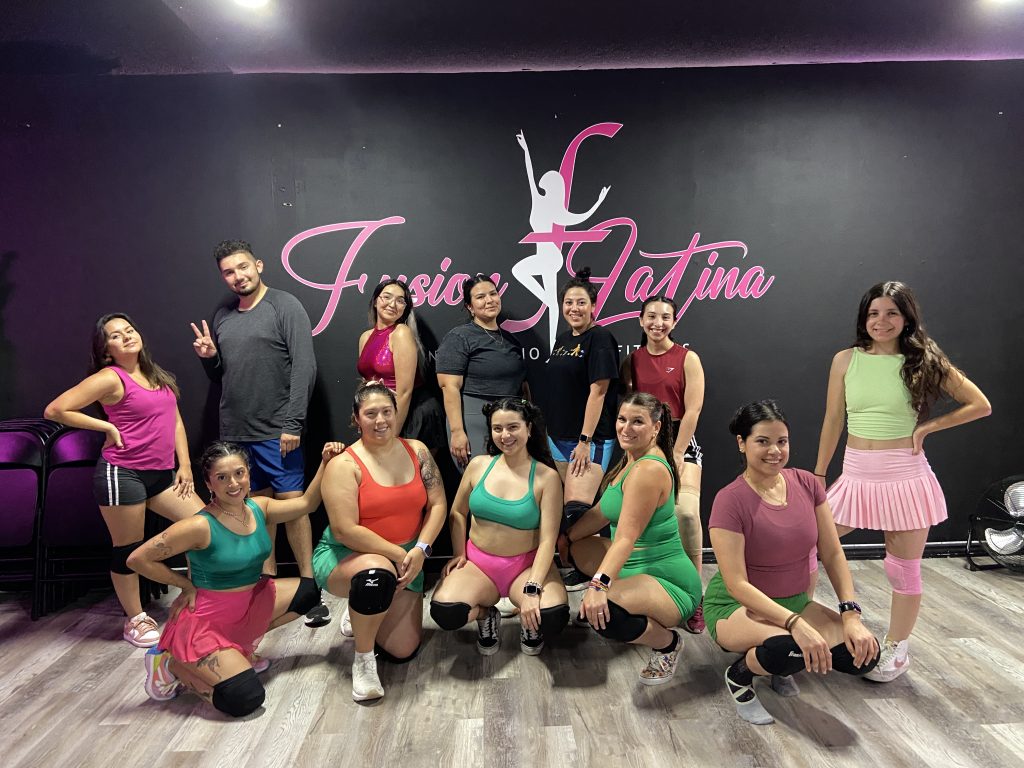 Twerk class Participants in a dance studio posing for a group photo behind a black wall that says Fusion Latina in pink script font.