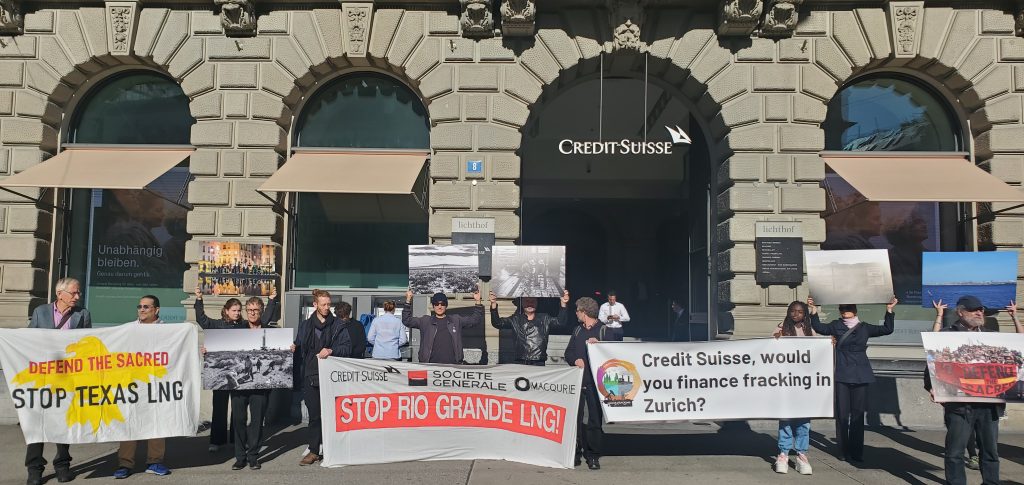 Protesters outside of a Swiss bank holding posters that say "stop rio grande LNG" in bright red letters and "stop Texas LNG" in black letters with a yellow eagle behind the letters.