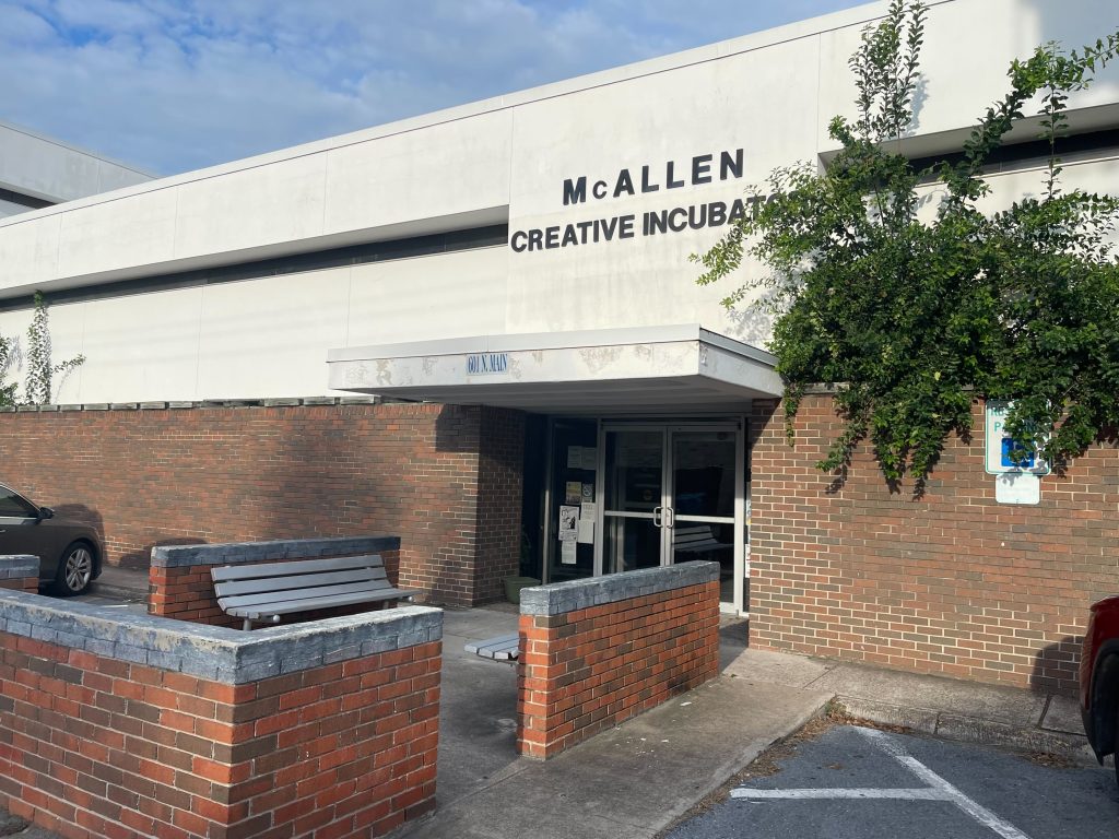 Outside view of a white building with black lettering that reads “McAllen Creative Incubator” with a red brick wall surrounding it.