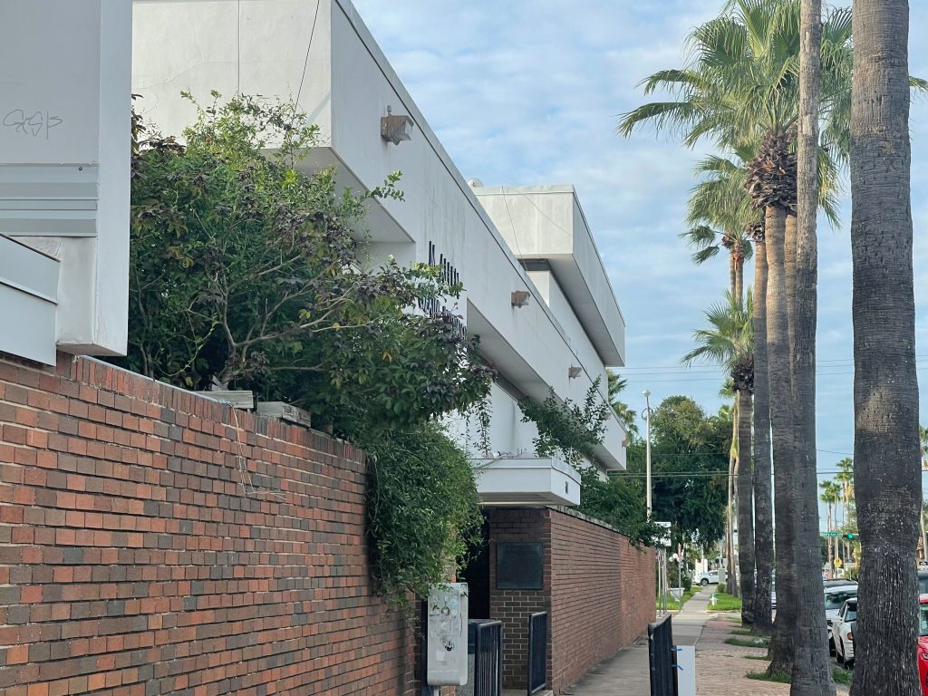 A sidewalk with a white building surrounded by red brick wall to the left of it and palm trees on its right.