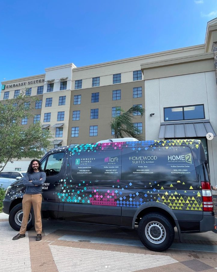 A Latinx person proudly standing next to his design on a black van.