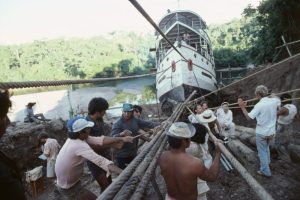 Multiple men holding thick ropes attached to a steamship, trying to move it up a steep hill.
