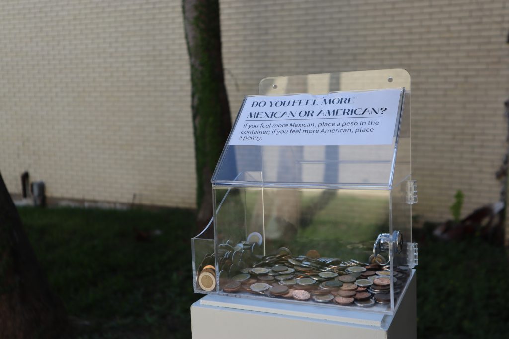 A transparent donation box is placed on top of a stand with a title that says, “Do you feel more Mexican or American?” The box beckons the viewer to place a peso or a penny into it based on their response.