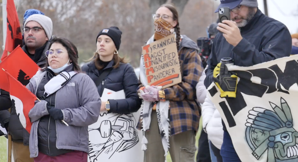 People at a rally standing, listening, and holding various signs.