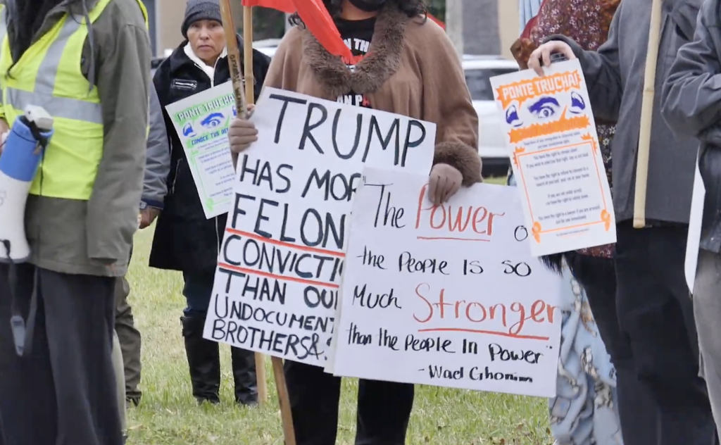 A person in a brown coat holding two signs at a rally.