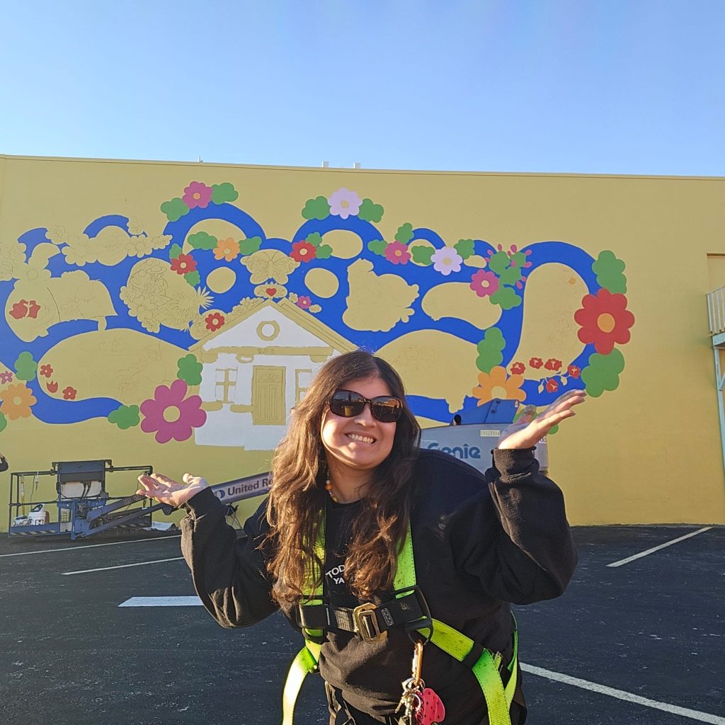 A woman smiling as she poses in front of a dandelion-colored mural.