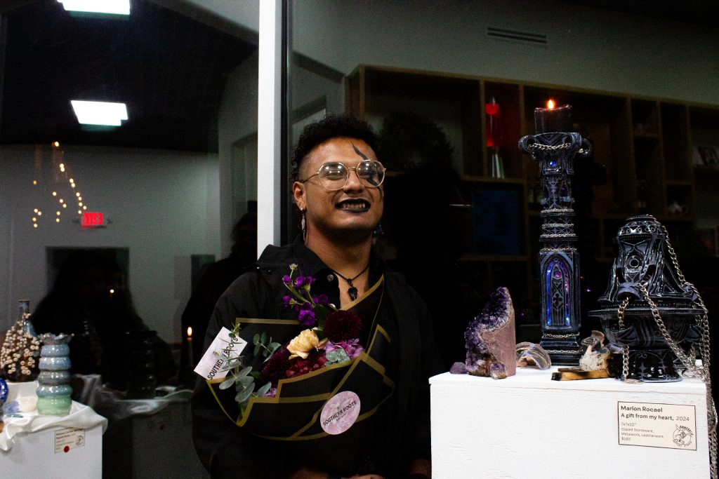 A person with curly hair smiling and holding flowers as they stand next to their Gothic-inspired ceramic pieces.