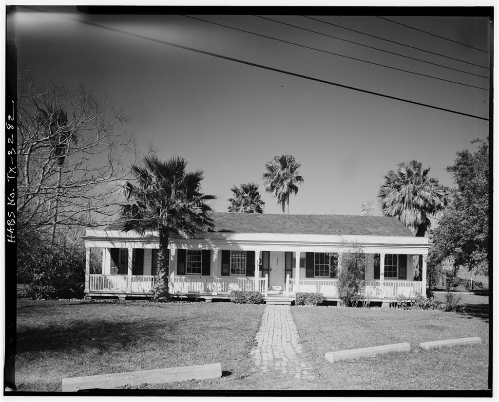 Black and white photograph of a house surrounded by palm trees and a walking path made of bricks leading to the front porch.