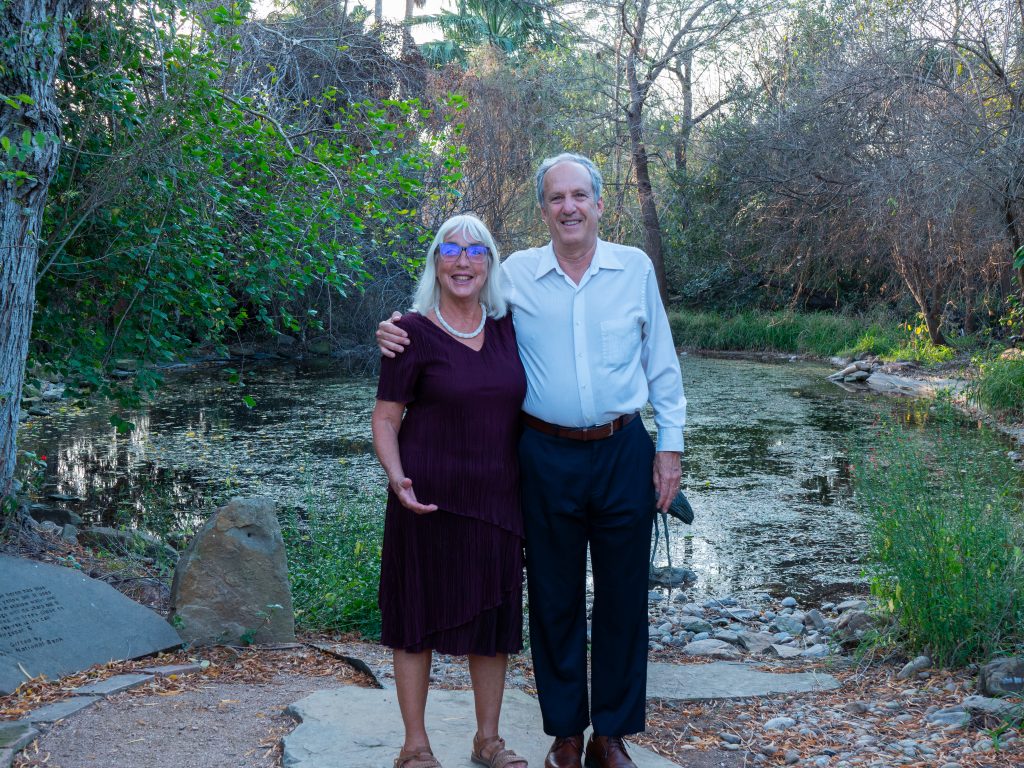 An elderly couple smiling, with white hear wearing dressy clothes, standing in front of a natural pond, surrounded by trees.