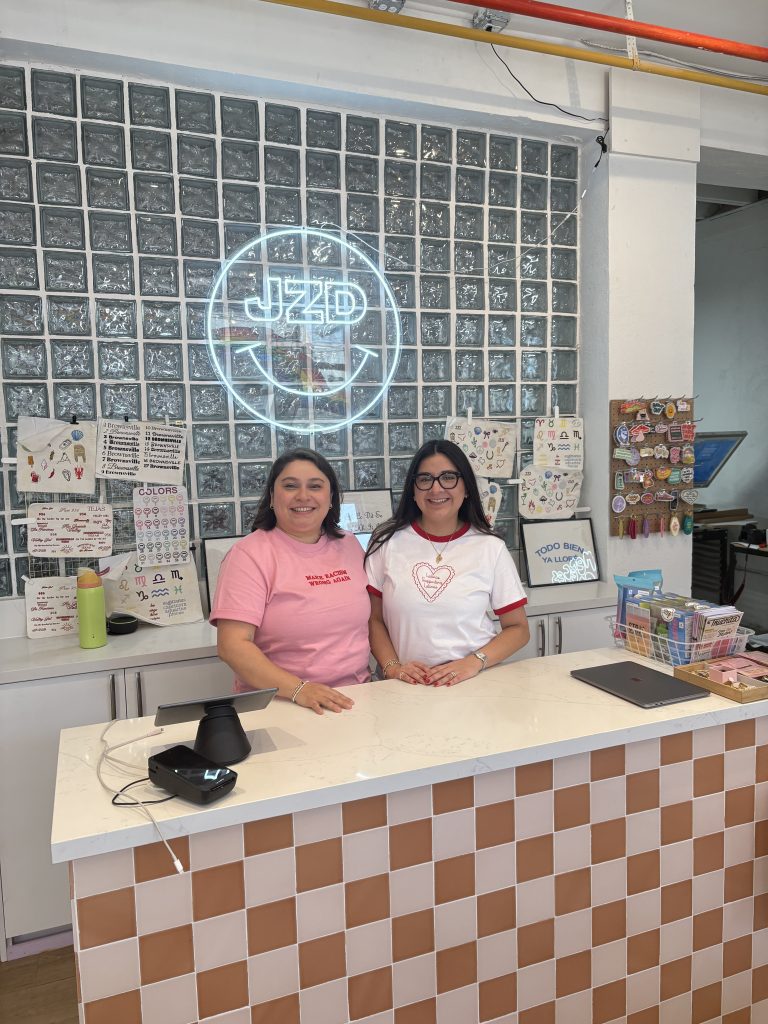 Two women behind a counter with a neon sign that reads "JZD."