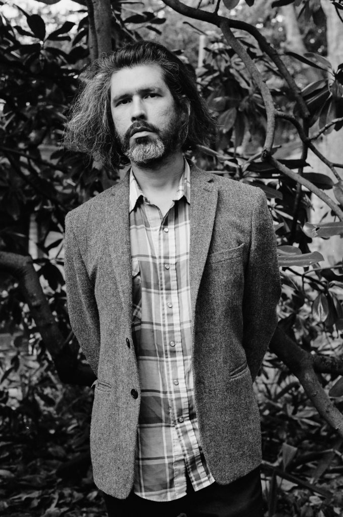 A black and white headshot of a man standing, posing in a stoic way.