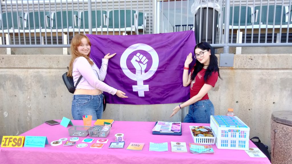 Two people posing behind a table with stickers, Plan B and informative pamphlets.