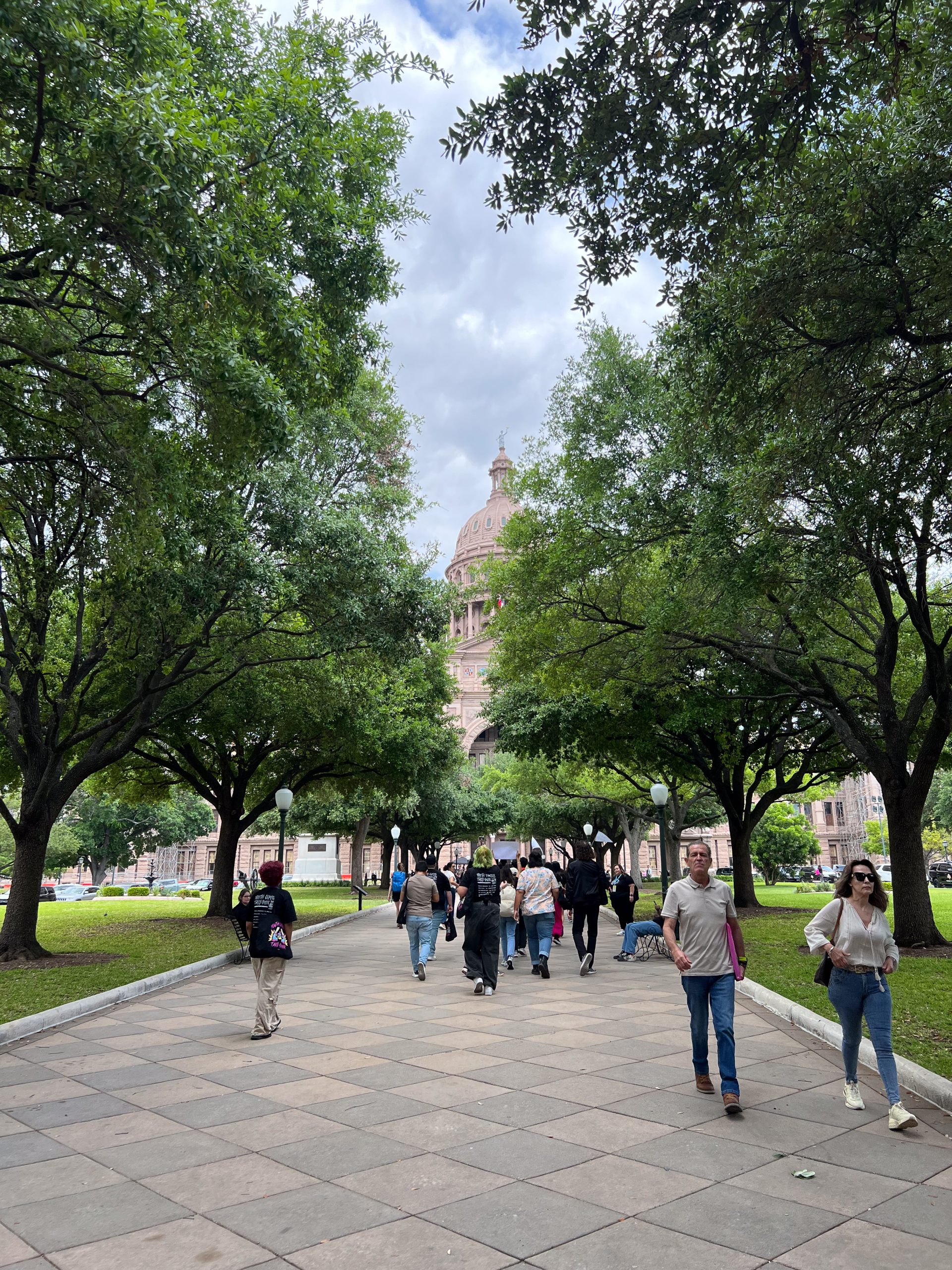 People walking to and from the Texas Capitol, seen covered by lush green trees.