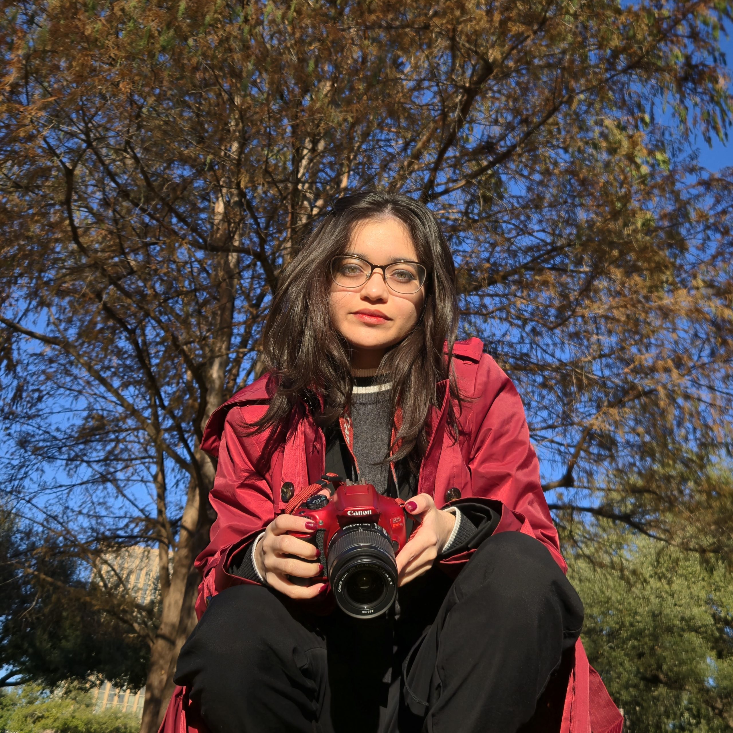 A photo of a woman holding a camera, a tree, and a building behind her. Photo Courtesy of Venisha Colón.