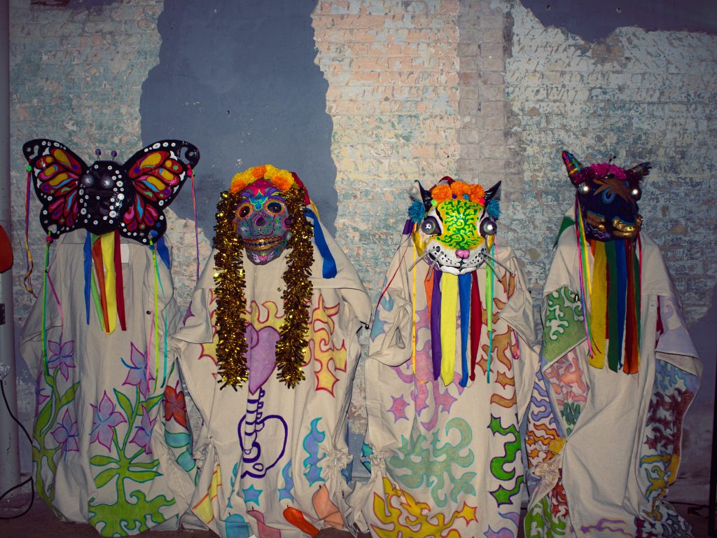 Four costumes with masks of various colors stand in front of a brick wall.