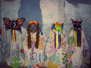 Four costumes with masks of various colors stand in front of a brick wall.