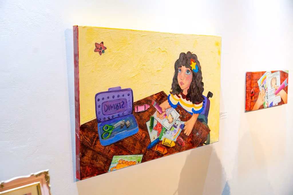 A painting of a girl in a wheelchair who is drawing on paper on a table.