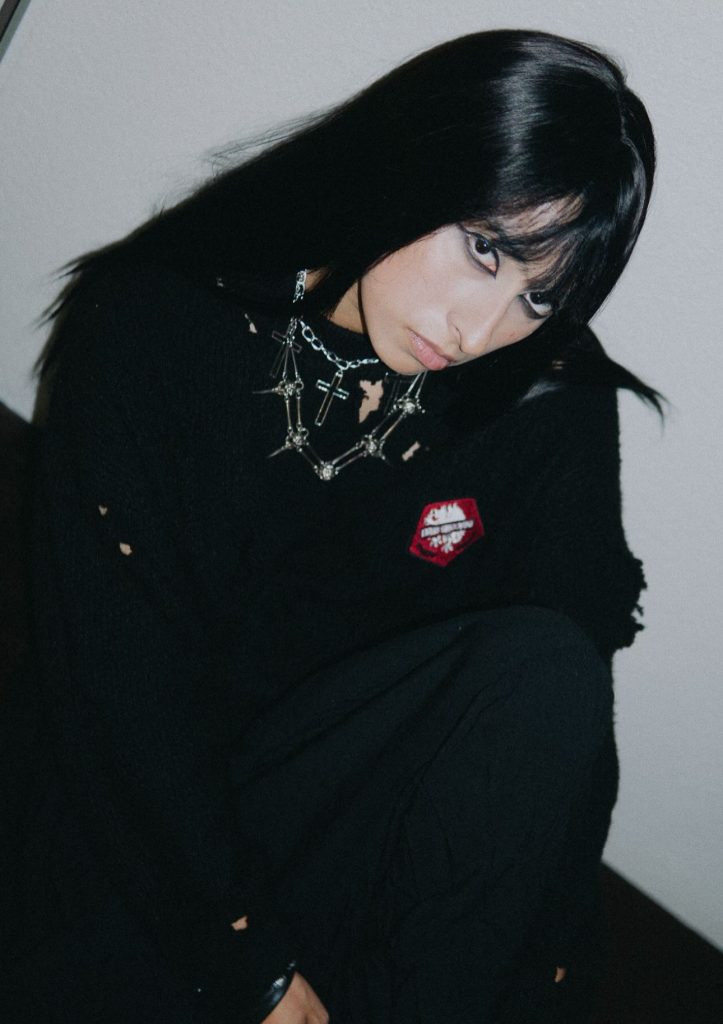 A young woman wearing dark clothes and eyeliner looks into the camera.