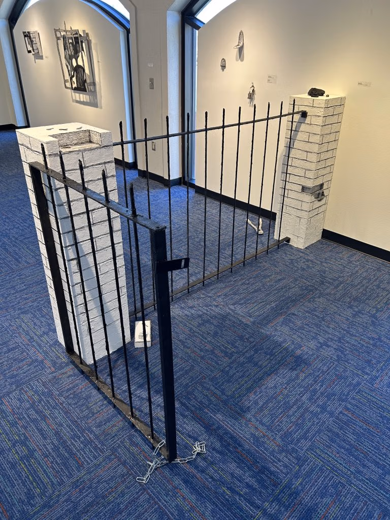An installation contains two sculpted brick pier columns supporting a burglar-bar gate that interrupts the gallery space. Several keys lie on the pier columns and on the floor.