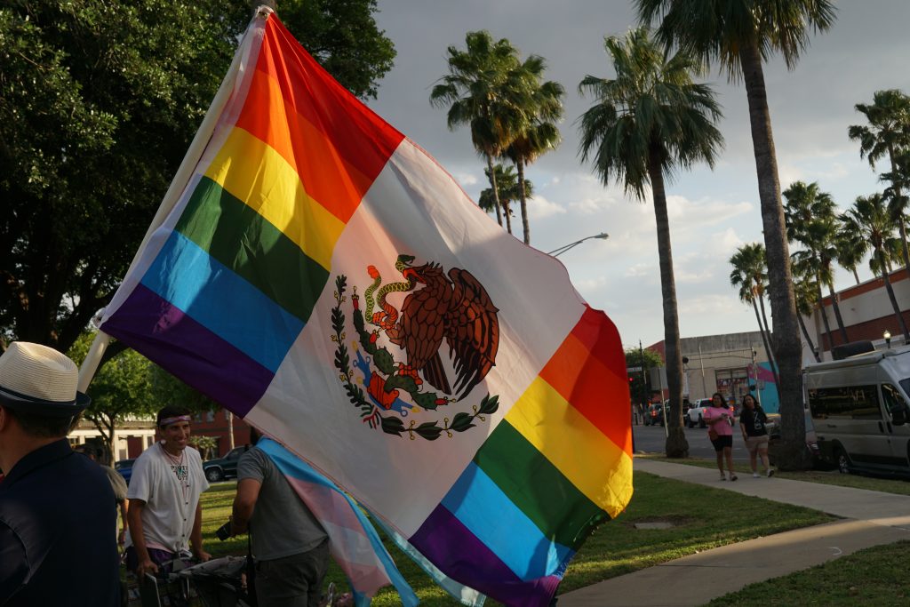 An LGBTQ+ flag with the symbol from the Mexican flag is waved in front of palm trees in downtown McAllen.