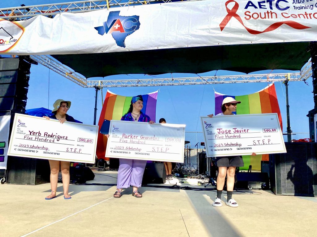 Three scholarship recipients each hold a big check for $500 and stand before two big Pride rainbow flags during RGV Pride at SPI in 2023.
